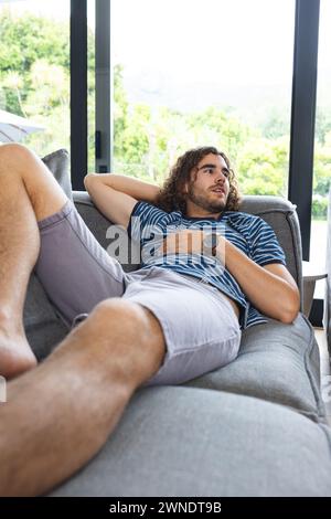 Young Caucasian man with curly hair lounges on a sofa, gazing thoughtfully with copy space Stock Photo