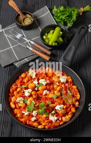 gigantes plaki, greek giant baked beans in a chunky tomato sauce sprinkled with feta cheese and fresh cilantro on skillet on black wooden table, verti Stock Photo