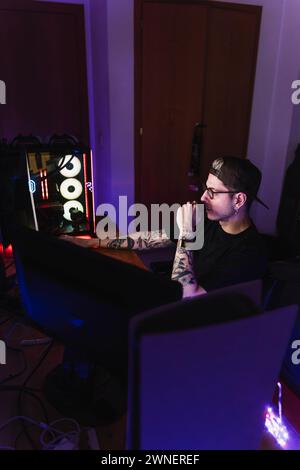 Side view of a concentrated young gamer wearing glasses using computer for playing games at home. Stock Photo