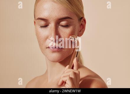 Young woman with closed eyes using face massager. Female with smooth freckled skin using face roller against a beige background. Young woman with clos Stock Photo