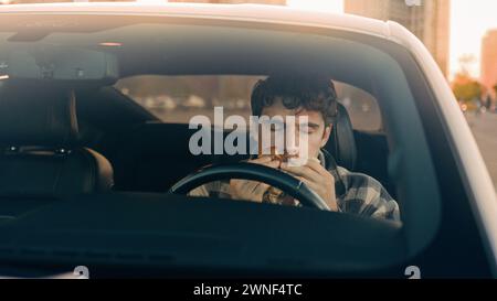 Caucasian man driver businessman eating cheeseburger inside auto in city parking lot. Hungry male guy eat hamburger junk fast food takeaway service in Stock Photo