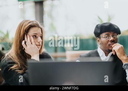 A focused young woman on a phone call and a contemplative man at a modern cafe engage in collaborative business work. Stock Photo
