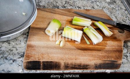 A selection of fresh leeks and a clove of garlic are laid out on a wooden cutting board, ready to be chopped with a nearby kitchen knife Stock Photo