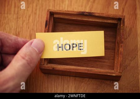 A man holding a piece of yellow paper with the word Hope on it in his hand over a small empty wooden box. Close up. Stock Photo