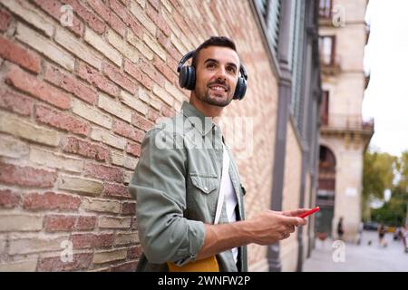Young handsome man smiling with wireless headphones holding cell phone leaning against brick wall. Stock Photo