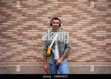 Portrait of stylish man looking happy at camera with wireless headphones leans against brick wall. Stock Photo