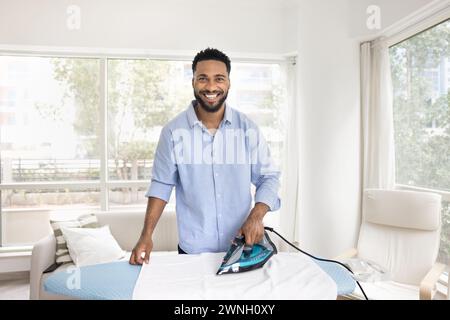 Cheerful attractive young African American homeowner man ironing linen Stock Photo