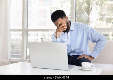 Exhausted frustrated African remote employee man sitting at laptop Stock Photo