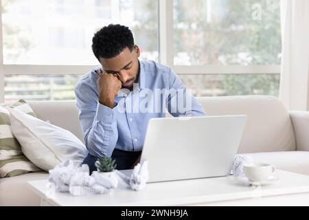 Tired sleepy African freelancer man sitting on sofa at laptop Stock Photo