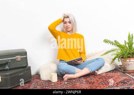 A casual woman with a tablet pauses thoughtfully, her hand in her hair, against a simple home backdrop with a vintage suitcase and lush greenery, refl Stock Photo