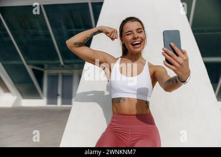 Sporty smiling young woman after running making selfie outdoors and showing biceps Stock Photo