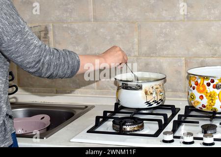 A woman stirs food with a spoon, which is cooked in a pan on a gas stove. Stock Photo