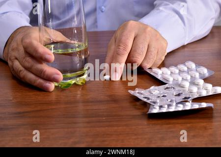 The picture shows a man who holds a glass of water in one hand, and the other takes a pill lying on the table. Stock Photo