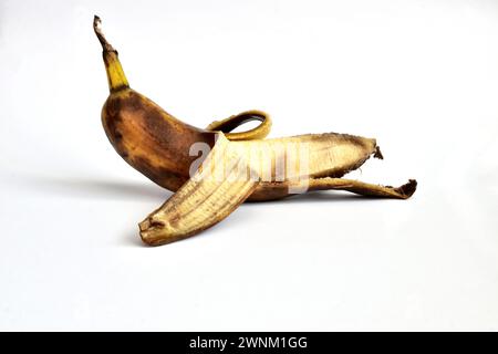 On a white background lies a banana that has not been completely peeled. The banana peel has dark spots. The banana is overripe. Stock Photo