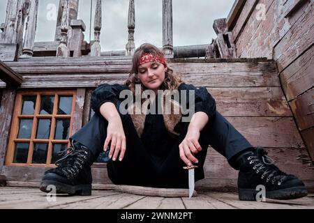 Corsair girl in a black clothes sits on the wooden floor of an retro pirate ship and draws a knife on the deck. Stock Photo