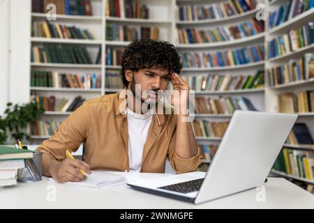 A focused young man is immersed in his studies at the library, surrounded by books, with a laptop for assistance. He seems to be preparing for exams, reflecting the life of a university or academy student. Stock Photo