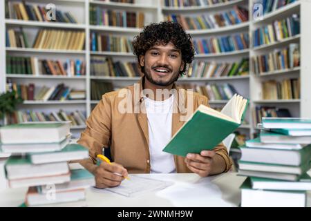 A happy young graduate student preparing for exams in a university library. Surrounded by large stacks of books, he represents dedication to academic success and the pursuit of knowledge in an institute of higher education. Stock Photo