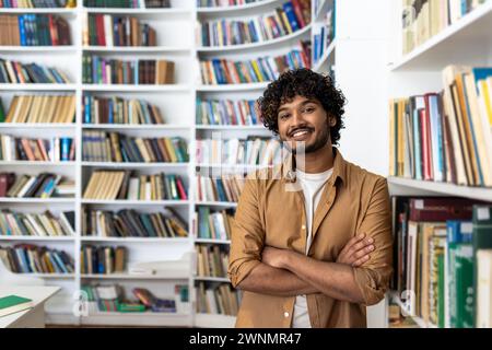 A cheerful young man with curly hair stands confidently with his arms crossed in a well-stocked library. He projects a vibe of intelligence and positivity amidst a collection of books. Stock Photo