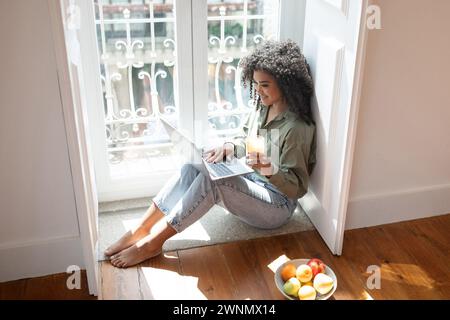 Happy young woman using laptop computer enjoying juice at home Stock Photo