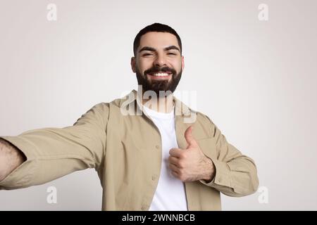 Cheerful bearded man taking a selfie with one hand while giving a thumbs up with the other Stock Photo