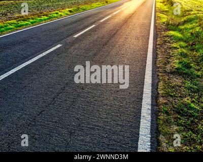 Sunlight casts shadows on an empty road surrounded by grass. Stock Photo