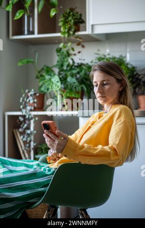 Young woman spends relaxing weekend room with houseplants, scrolling through news feed smartphone. Stock Photo
