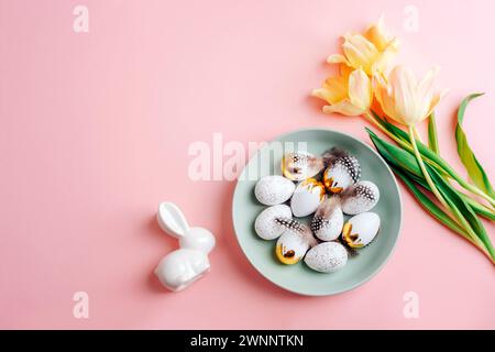 Easter layout with plate of Easter eggs, yellow tulips and bunny on pink table. Top view, flat lay, copy space. Stock Photo