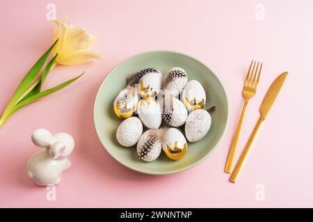 Table place setting with green plate with Easter eggs, yellow tulip and bunny on pink background. Top view, flat lay. Stock Photo