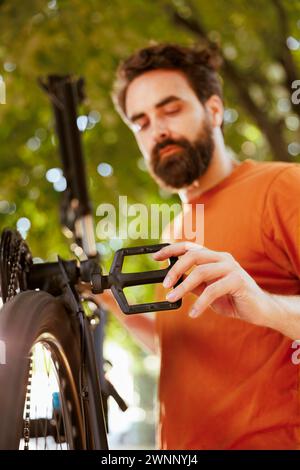 Sports-loving dedicated man repairing bicycle parts using professional tools in home yard. Healthy caucasian male cyclist assessing and mending damaged bicycle as summer activity. Stock Photo