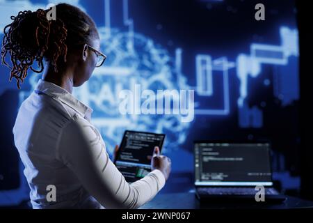 BIPOC engineer updating neural networks made up of interconnected nodes, writing intricate binary code scripts on laptop and tablet. Computer operator using programming to upgrade AI simulation model Stock Photo