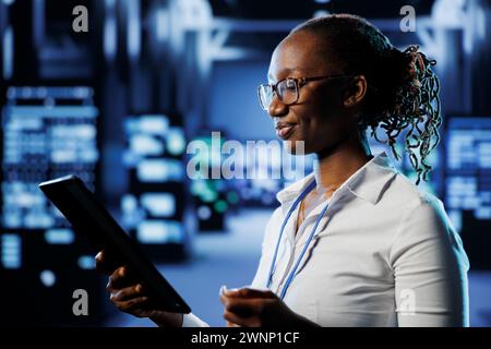 Upbeat admin in data center uses tablet to prevent system infrastructure overload. Precise expert between blade servers ensuring enough network bandwidth for smooth operations Stock Photo