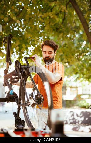 Healthy sports-loving man securing bike wheel for outdoor leisure cycling. Active young caucasian male cyclist doing yearly maintenance and adjusting bicycle tire in yard during the summer. Stock Photo