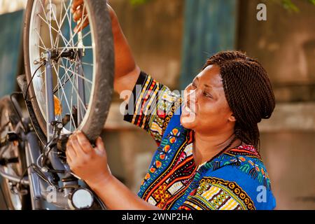 Active young woman performing yearly maintenance and adjusting bicycle tire in yard during the summer. Healthy sportloving african american lady securing bike wheel for outdoor leisure cycling. Stock Photo