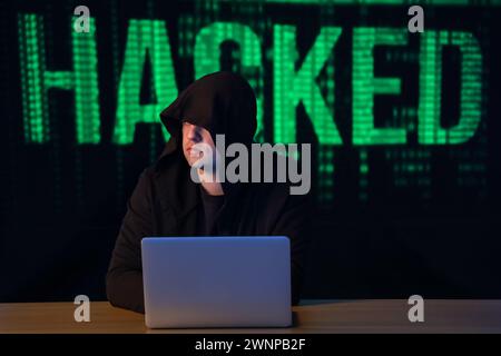 Hacker with laptop at table against word HACKED on dark background Stock Photo