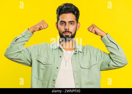 I am strong and independent. Young Indian man showing biceps and looking confident, feeling power strength to fight for rights, energy to gain success win. Guy isolated on yellow studio background Stock Photo