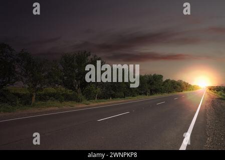 Empty asphalt road and trees under dark sky with rising sun Stock Photo