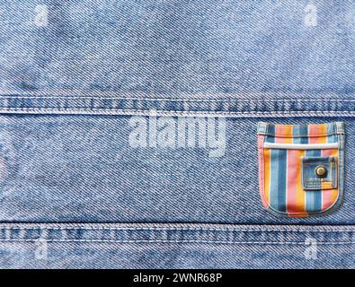 Grey and light grey denim background with a seam and colourful pocket. Light gray color denim jeans fabric texture and striped multicolor pouch. Copy Stock Photo