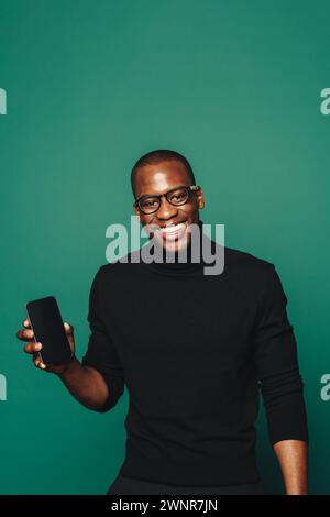 Stylishly dressed in casual clothing, a young man holds a trendy mobile phone against a vivid green background. With confident and happy demeanor, he Stock Photo