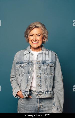 Portrait of a confident and cheerful mature woman with grey hair standing in a studio. She is wearing a fashionable denim outfit and a casual blue bac Stock Photo