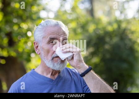 Close-up portrait of a sick elderly man with a beard standing in a park in nature, wiping his nose with a napkin, suffering from a runny nose and seasonal allergies. Stock Photo