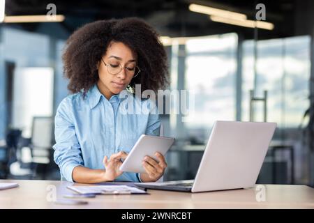 Concentrated and serious African American woman in blue shirt and glasses sitting in modern office at desk with laptop and using tablet. Stock Photo
