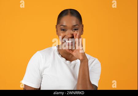 Cheerful African American woman stands against orange backdrop sharing secret Stock Photo
