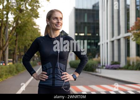 A fit and happy woman takes a break from running, her hands on her hips as she pauses to enjoy the city scenery. Earphones in, she's dressed in athletic gear, ready to continue her fitness routine. Stock Photo