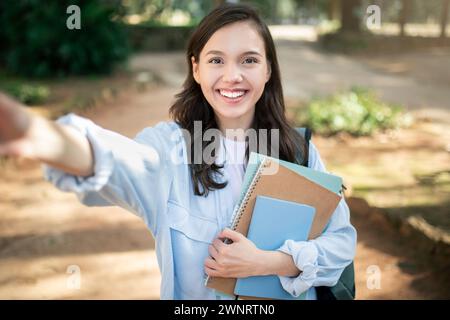 A radiant young woman smiles at the camera, taking a selfie while clutching notebooks on a sunny outdoor campus Stock Photo