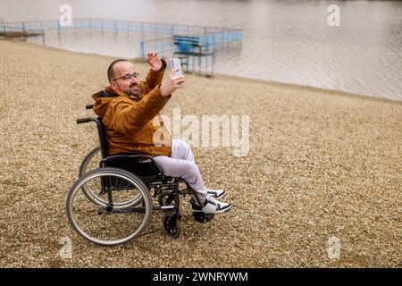Paraplegic handicapped man in wheelchair is taking selfie with smartphone outdoor. Stock Photo