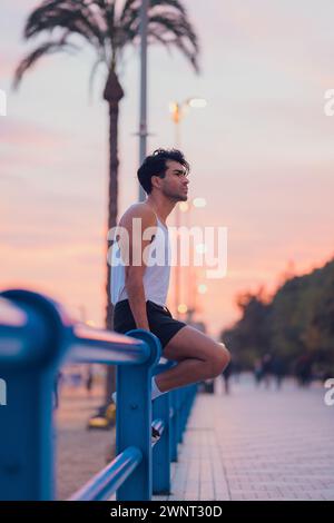Pensive male athlete resting on a blue railing at beachside at sunset Stock Photo