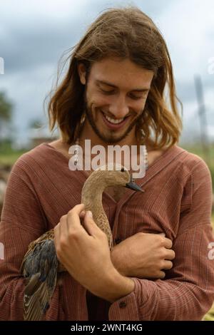 Smiling long-haired man holds duck in his hands. Stock Photo