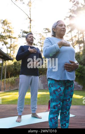 Senior biracial woman and man practice yoga outdoors, standing on mats at home Stock Photo
