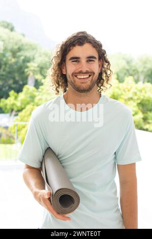 Young Caucasian man with curly hair holds a yoga mat, smiling outdoors Stock Photo