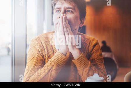 Lonely man sitting inside, deep in thought, hands pressed together near face, looking through window. Stock Photo